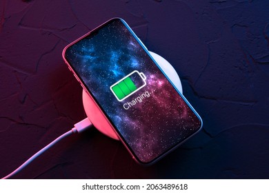 Mobile smart phone on wireless charging device on dark neon red and blue color background. Icon battery and charging progress lighting on screen.smartphones connected to power source.Low battery.