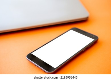 Mobile Technology. Digital Mockup. Online Connection. Smartphone With White Blank Screen And Closed Laptop Isolated Orange.