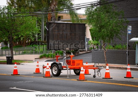 A mobile traffic control device displays a message about a road closure, surrounded by safety cones on a street