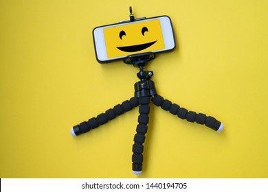 Mobile Tripod Isolated On Yellow Background. Cell Phone On A Tripod. Cheerful Smile On The Cell Phone Screen