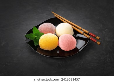Mochi dessert on a black background in a black plate with mint leaves. There are bamboo sticks nearby. View from above. Perfect retouching.