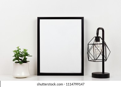 Mock up black frame, succulent plant and industrial style lamp on a shelf or desk. White shelf and wall. Portrait frame orientation. - Powered by Shutterstock