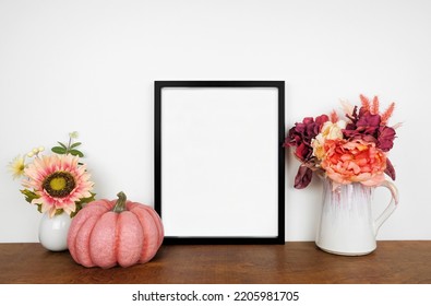 Mock Up Black Picture Frame With Pink Hue Autumn Flowers And Pumpkin On A Wood Shelf Against A White Wall. Fall Concept. Copy Space.