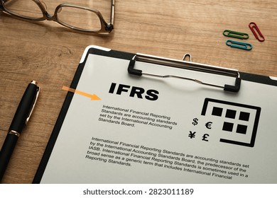 Mock IFRS document displayed on wooden desk with business accessories, featuring currency symbols and accounting standards information. Perfect for finance articles, compliance education content, and corporate blog illustrations.