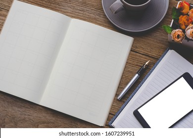 Mock up image of black mobile smart phone with blank white screen, weekly planner notebook, pen, cup of coffee and flowers on wooden table background. 