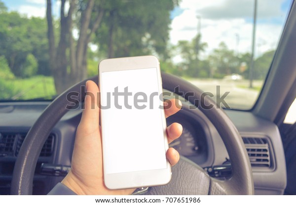 Mock up image of a hand holding a smart phone\
while driving with blank white screen. Technology. Spinning wheel\
in the background.
