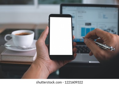 Mock Up Image Of Man Hand Holding Blank Screen Mobile Smart Phone With Laptop Computer And Cup Of Coffee On Desk At Office, Over Shoulder View