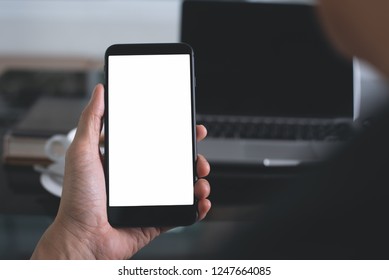 Mock Up Image Of Man Hand Holding Blank Screen Mobile Smart Phone With Laptop Computer And Cup Of Coffee On Desk At Office, Over Shoulder View, Toned Image