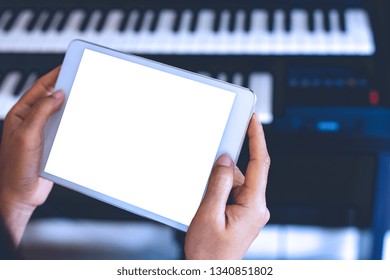 Mock Up Image Of Woman Hands Holding And Using Digital Tablet With Blank White Screen With Piano Blurred Background In Music Room. Clipping Path And Copy Space For Text And Advertising.