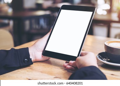Mock up image of a woman's hands holding black tablet pc with white blank screen and coffee cup on wooden table in cafe background