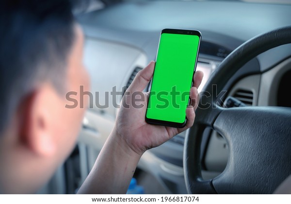 Mock up of man using mobile smart phone inside\
a car. Driver hand holding blank green screen smartphone, searching\
address and pin location via map navigator application,\
transportation technology