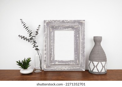 Mock up rustic gray weathered frame with succulent plant, eucalyptus branch and clay vase on a shelf or table. Wood shelf and white wall. Portrait frame orientation.