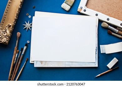 Mock up for watercolorists, illustrators, lettering, artists presentations. On a blue background a drawing tablet, a gilded picture frame, brushes, a pencil and white sheets of A4 paper. Top view - Powered by Shutterstock