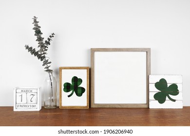Mock up wood frame with St Patricks Day decor on a wood shelf. Shabby chic wood signs, calendar and green branches. Square frame against a white wall. Copy space.