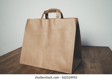 Mock-up Of Big Brown Paper Package With Handles, Blank Craft Shopping Bag With Area For Your Logo Or Design, White Wall In The Background, Food Delivery Concept