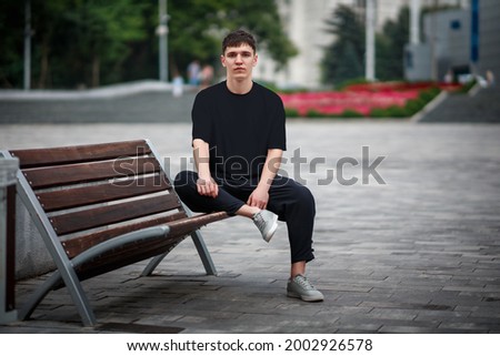 Mockup of a black casual t-shirt on a guy sitting on a blurred background of a park, on a bench, front view, for design presentation. Blank clothing template for online store advertising.Urban style