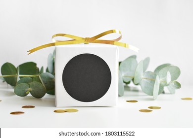 A Mock-up Of A Black Round Sticker On A Festive White Box With A Gold Ribbon Surrounded By Eucalyptus Branches And Gold Confetti.