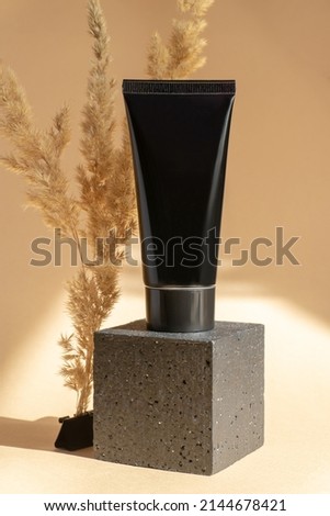 Mockup of black tube for cosmetics on geometric 3d podium. Plastic container for cream, lotion, toothpaste on square stand, surrounded by dry blade of grass. Beige background, vertical image.