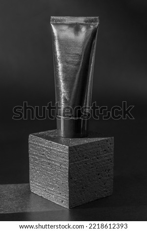 Mockup of black tube of cosmetics on square platform. Unmarked container for cream, lotion, toothpaste on 3d podium. Vertical image, close-up, black background with sun glare.