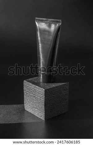Mockup of black tube of cosmetics on 3d podium. Unmarked container for cream, lotion, toothpaste on square platform. Vertical image, close-up, black background with sun glare,