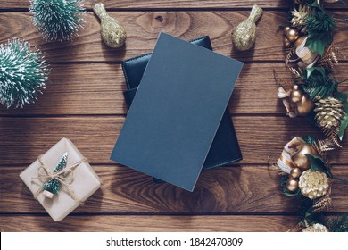 Mockup Blank Black Book Cover With Christmas Gift Box, Xmas Ornaments And Christmas Tree Model Decor On Wooden Table Background. Flat Lay, Top View With Copy Space