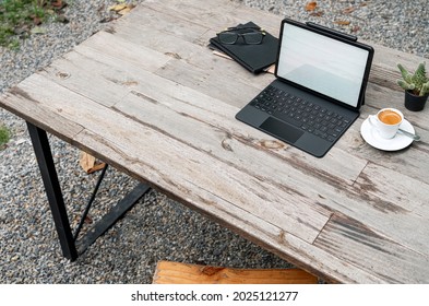 Mockup blank screen tablet with magic keyboard, coffee cup and notebook on wooden table outdoors.