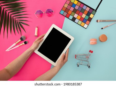 Mockup With Blank Tablet Screen In Female Hands And Various Beauty Cosmetics, Eyeshadow Palette, Brushes, Sunglasses On Blue And Pink Background, Top View