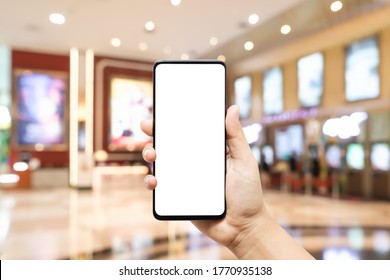 Mockup Blank White Screen Mobile Phone Hand Holding Smartphone  With Blurred Image Hall Of Ticket Sales Counter At Movie Theater. Background Concept For Buy Cinema Tickets, Concerts, Etc