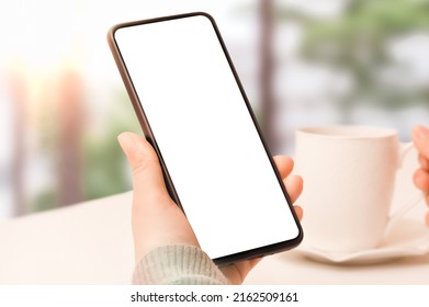 Mockup Cellphone. Close Up View Of Woman Hand Holding Mock Up Smart Phone With Blank Screen And Cup Of Coffee. In Cafe At White Table Against Window With Summer Landscape. Hand Holding Mobile Phone