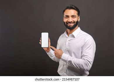 Mock-up concept. Handsome confident bearded Indian man in formal wear showing smartphone with empty screen, points finger at blank device display isolated on black, male advertising new mobile app