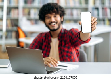 Mock-up And Copy-space Concept. Photo Of Defocused Indian Or Arabian Male Student, Sitting At Table In Library, Showing His Smart Phone With Blank White Screen For Advertising And Presentation