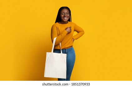 Mockup For Design. Cheerful African American Lady Holding Blank White Eco Tote Canvas Bag With Copy Space, Positive Black Female Posing Over Yellow Background In Studio, Smiling At Camera