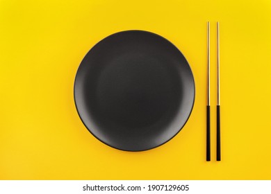 Mockup Empty Black Round Plate With Gilded Chinese Chopsticks Flat Lay On Yellow Background With Copy Space.