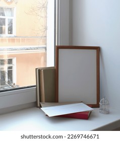 Mockup of empty photo frames or paintings, books, postal envelopes and a decorative cage on a white window sill against a window background.