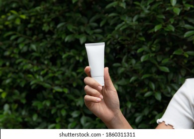 Mockup Facial Skincare White Tube Product With Blank Label Holding By Woman’s Hand With Ficus Annulata (Banyan-Baum) Green Bush Nature Background And Copy Space