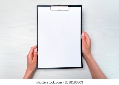 mockup female hands hold clipboard with a4 paper over light wooden table. top view close-up. flat lay.