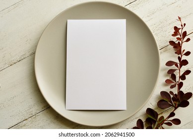 A mock-up of a gray plate with a white invitation card on a white wooden background with red branch of a plant close-up.