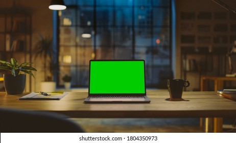 Mock-up Green Screen Laptop Standing On The Desk In The Modern Creative Office. In The Background Warm Evening Lighting And Open Space Studio With City Window View.