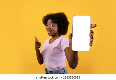 Mockup image of african american lady showing smartphone with blank screen over yellow studio background. Mobile phone with empty screen in female hand, closeup, selective focus