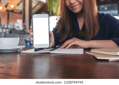 Mockup image of an asian woman holding and showing white mobile phone with blank desktop screen with coffee cups and notebooks on vintage wooden table in cafe