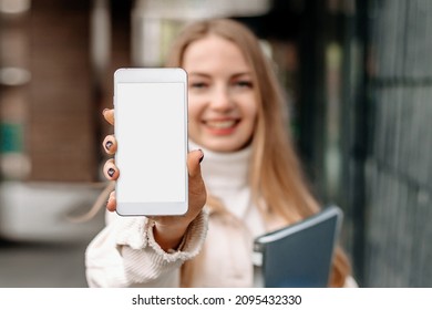Mockup Image Of A Blonde Female Student Showing A Mobile Phone With Blank White Screen Against The Background Of The University Building