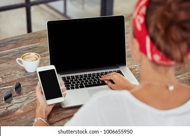 Mockup image of fashionable woman sits in front of opened laptop computer and holds modern cell phone with blank copy screen for your inforamtion or promotion, has breakfast in cozy restaurant