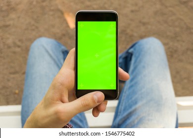 Mockup Image Of Hand Holding Black Mobile Phone With Blank Green Screen. Student On The Bench And Looks At The Vertically Positioned Smartphone Screen. Close Up.