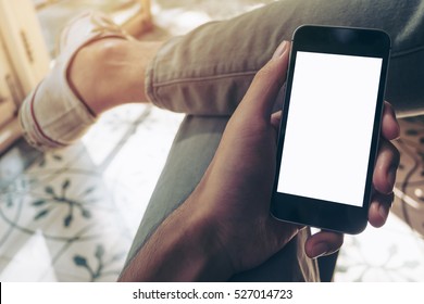 Mockup Image Of Hand Holding Black Mobile Phone With Blank White Screen On Thigh With White Canvas Shoes At Vintage Tile Floor In Cafe , Feeling Relax