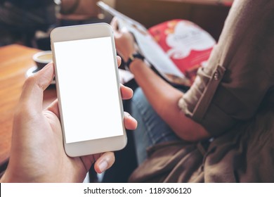 Mockup image of hand holding white mobile phone with blank screen with woman reading books in modern cafe - Powered by Shutterstock