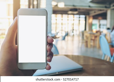 Mockup image of hand holding white mobile phone with blank white screen and silver laptop on vintage wood table in cafe