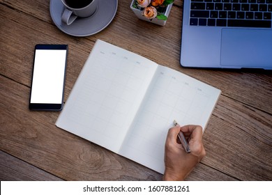 Mockup Image Of Hand Writing Note On Weekly Planner With Mobile Smart Phone With Blank White Screen And Laptop Computer On The Wooden Desk. Top View. Flat Lay. 