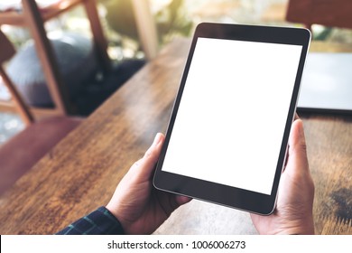 Mockup Image Of Hands Holding Black Tablet Pc With Blank Desktop White Screen On Wooden Table In Cafe Background