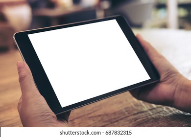 Mockup Image Of Hands Holding Black Tablet Pc With White Blank Screen On Wooden Table Background In Cafe