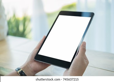 Mockup Image Of Hands Holding Black Tablet Pc With Blank White Screen On Wooden Table In Cafe